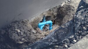Exposed blue plumbing pipes in a dirt trench near a building foundation.