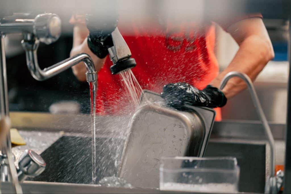 Hands washing a metal pan under running water with kitchen gloves.