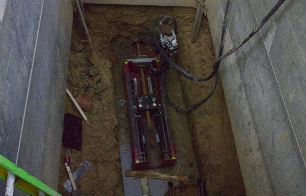 Construction site with drilling machinery in a deep trench and surrounding dirt.