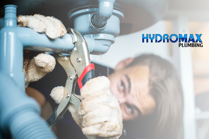 A plumber uses a wrench on a pipe under a sink, wearing gloves. The "Hydromax Plumbing" logo is on the right. The scene conveys focus and professionalism.