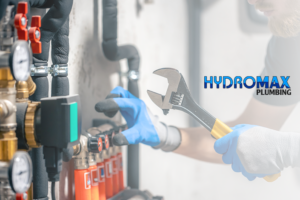 A plumber in blue gloves uses a wrench on pipes, surrounded by gauges and valves. The scene conveys precision and expertise. “Hydromax Plumbing” logo at bottom.